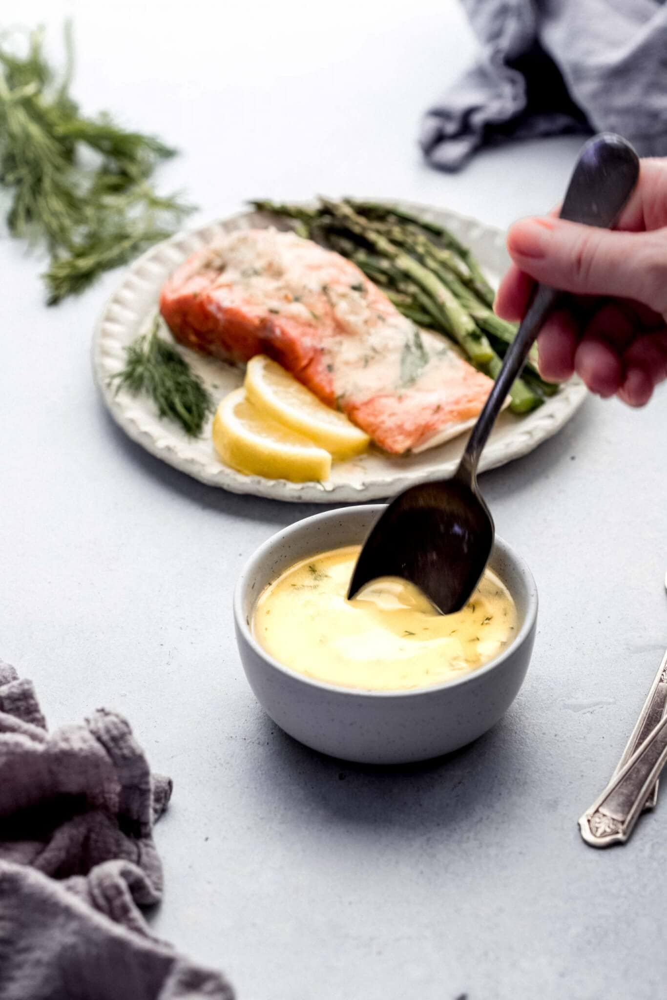 Spoon stirring lemon butter sauce in small white bowl next to plate of salmon and asparagus