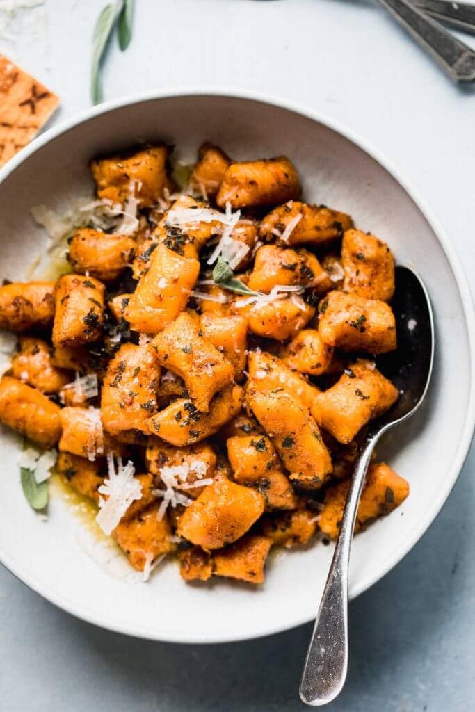 Sweet potato gnocchi in white bowl next to sage leaves and parmesan rind.