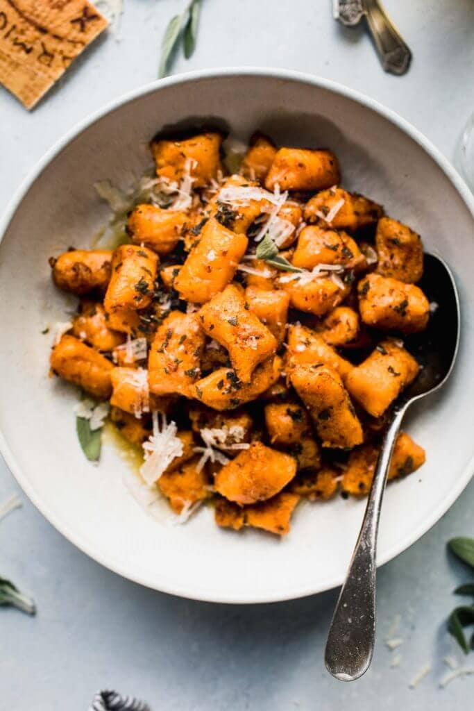 Sweet potato gnocchi in white bowl next to sage leaves and parmesan rind.