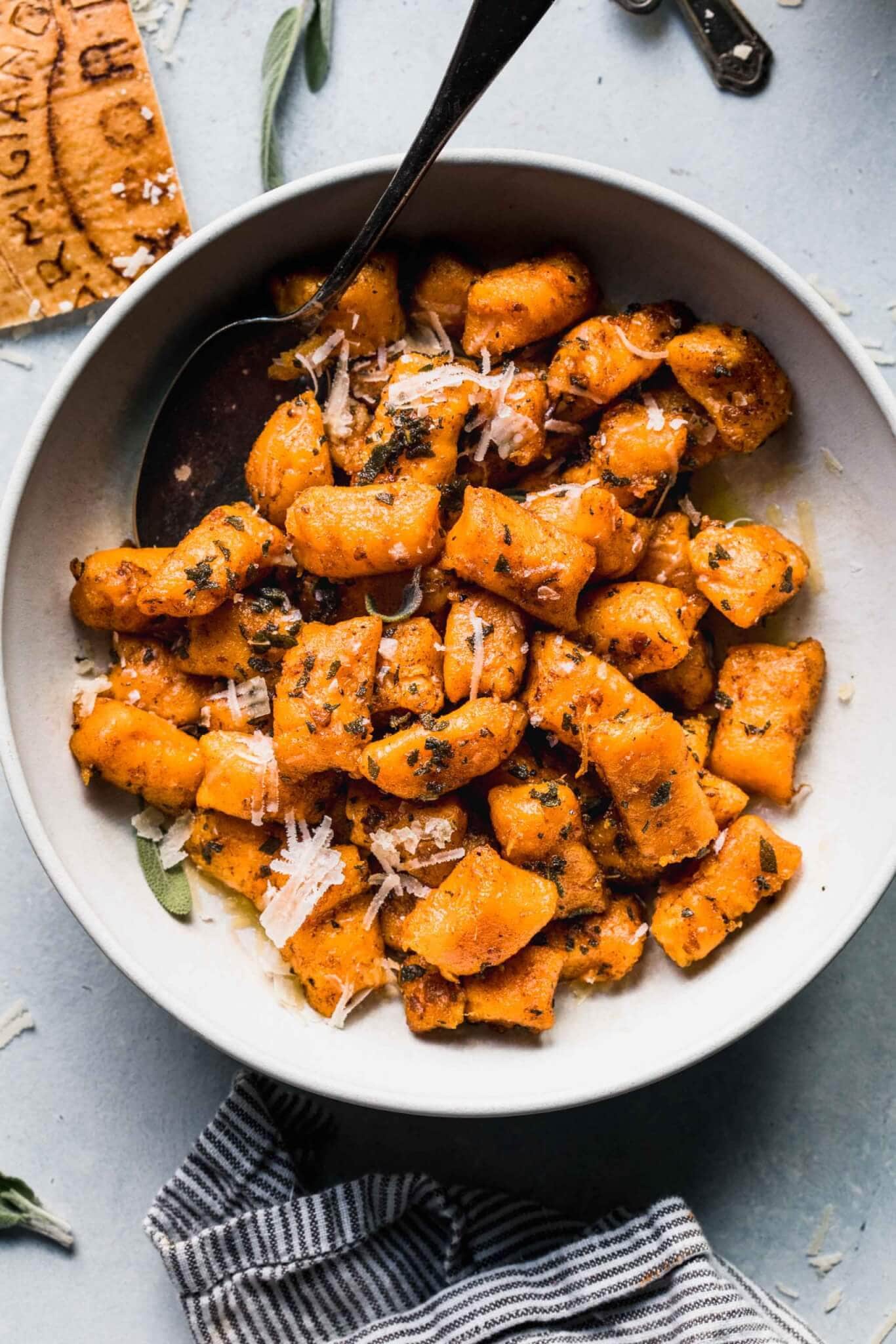 Sweet potato gnocchi in white bowl next to sage leaves and parmesan rind.