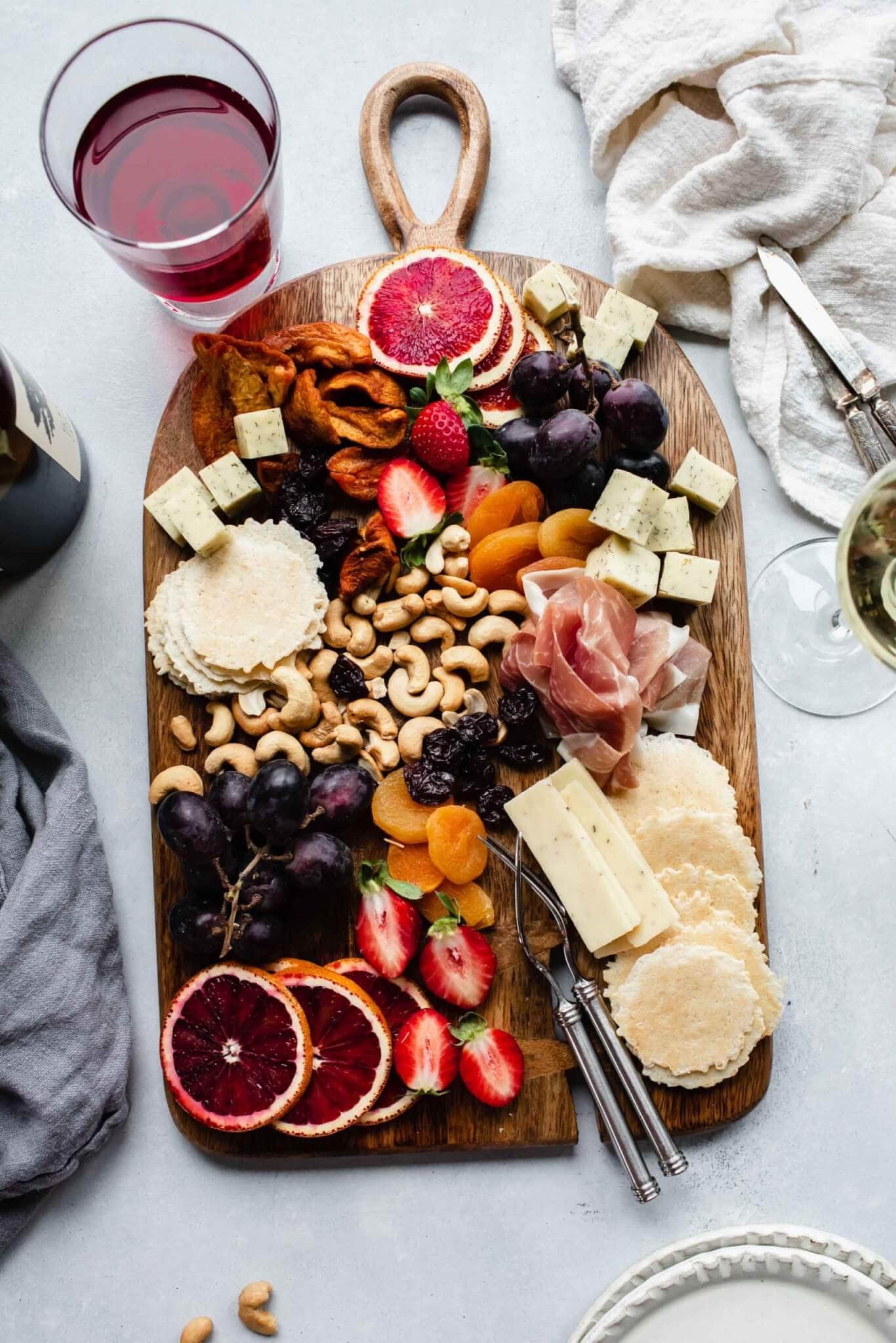 Cheeses, crackers and fruits and nuts on wood serving tray next to glass of wine.