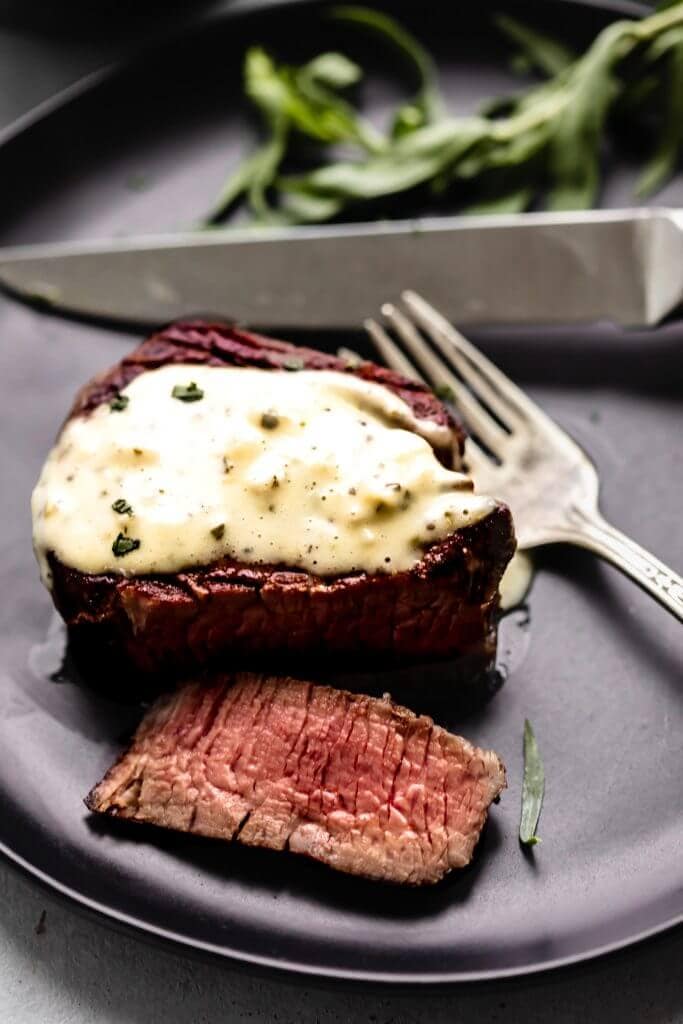 Steak with bearnaise sauce drizzled on top, on a black plate with fork and knife