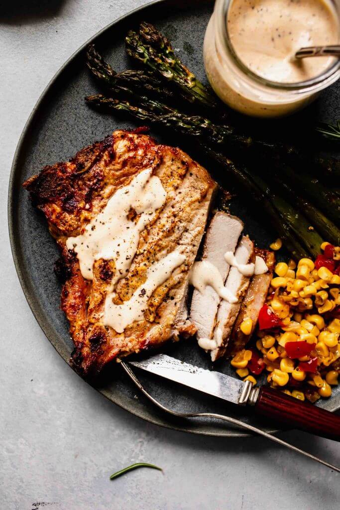 Sliced pork chop on grey plate next to sauteed corn and asparagus. 