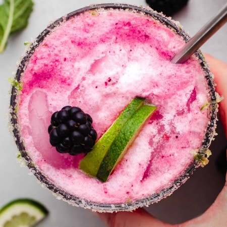 Hand holding blackberry mojito garnished with lime wedges and straw.
