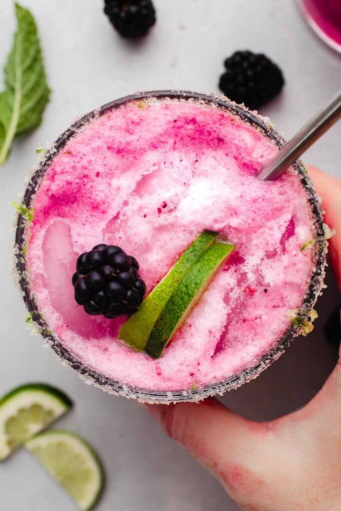 Hand holding blackberry mojito garnished with lime wedges and straw.