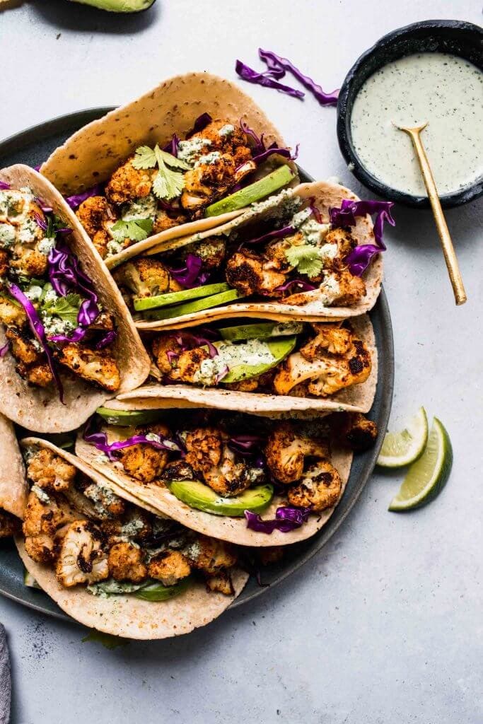 Cauliflower tacos arranged on grey plate next to bowl of crema.