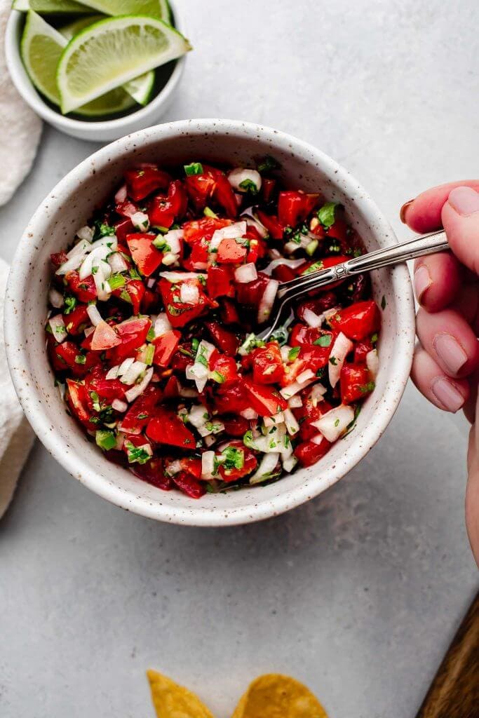 Hand dipping spoon into bowl of pico de gallo.