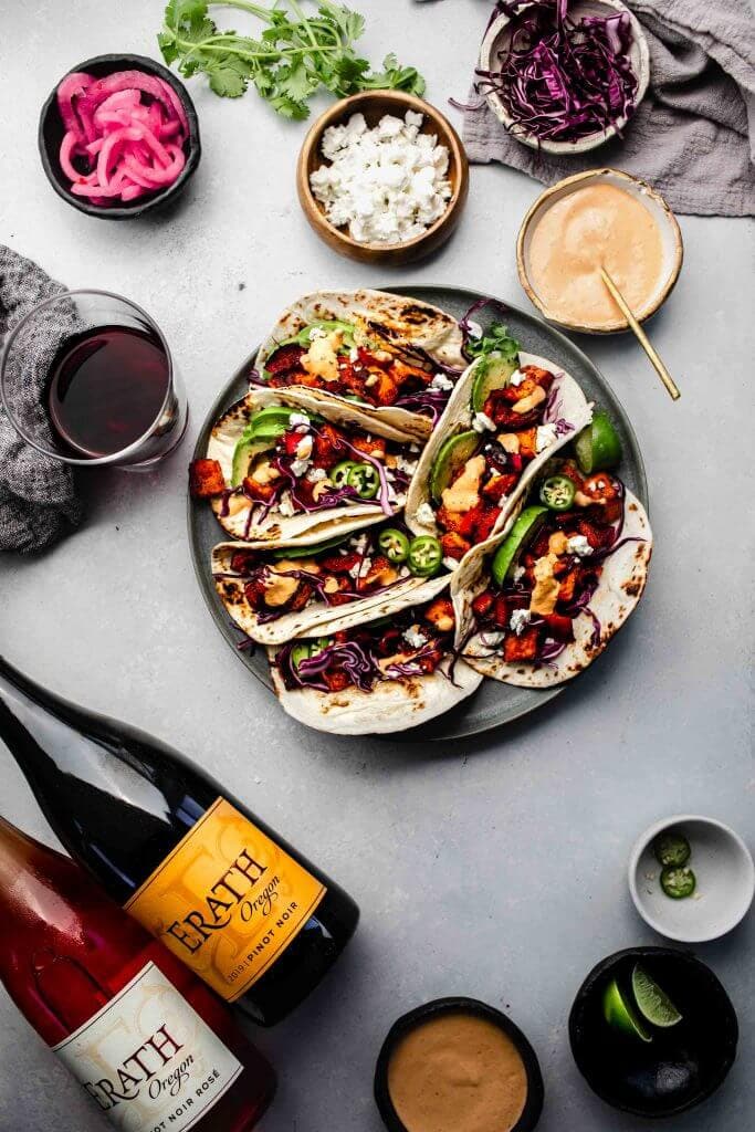 Tacos on grey plate next to bottles of wine and toppings. 