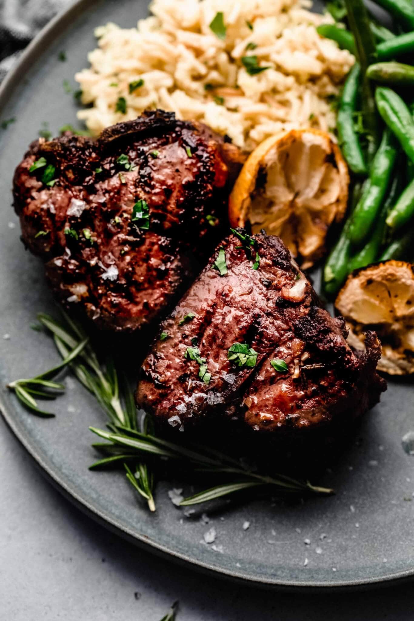 Two lamb chops on plate next to grilled lemons, green beans, and pilaf.