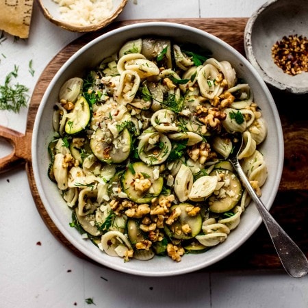 Pasta salad in bowl on wooden cutting board next to bowl of pepper flakes.