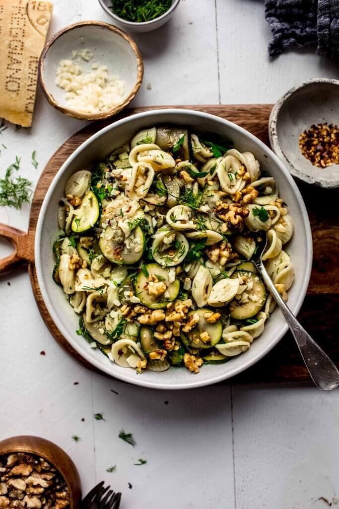 Pasta salad in bowl on wooden cutting board next to bowl of pepper flakes.