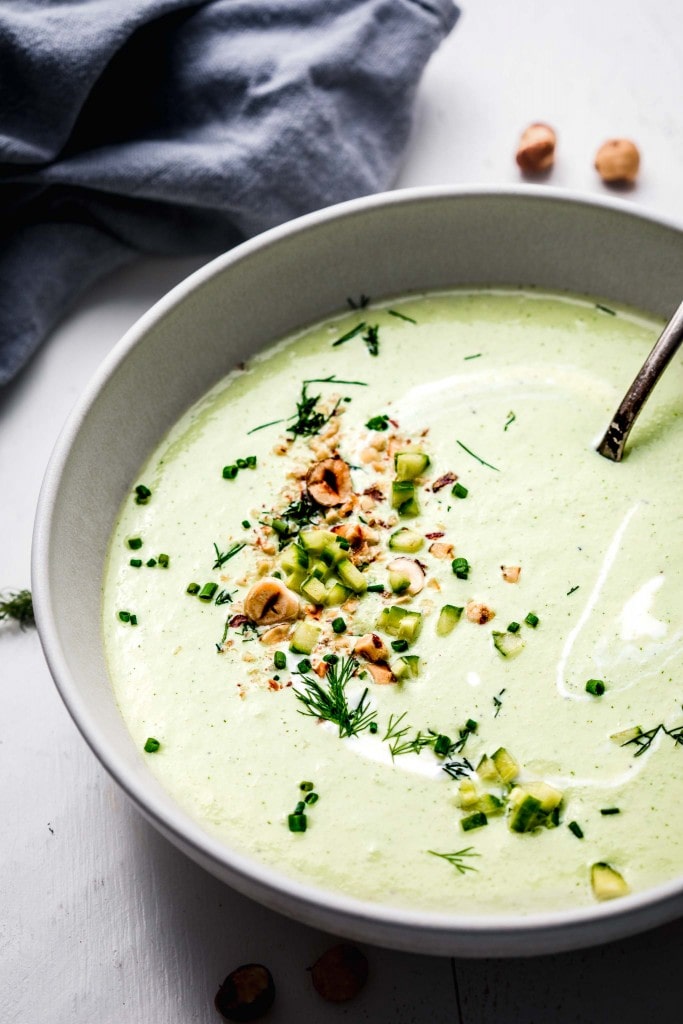 Side view of bowl of chilled cucumber soup with spoon and topped with hazelnuts. 