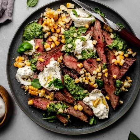 Grilled flank steak arranged on plate topped with corn, pesto and burrata cheese.