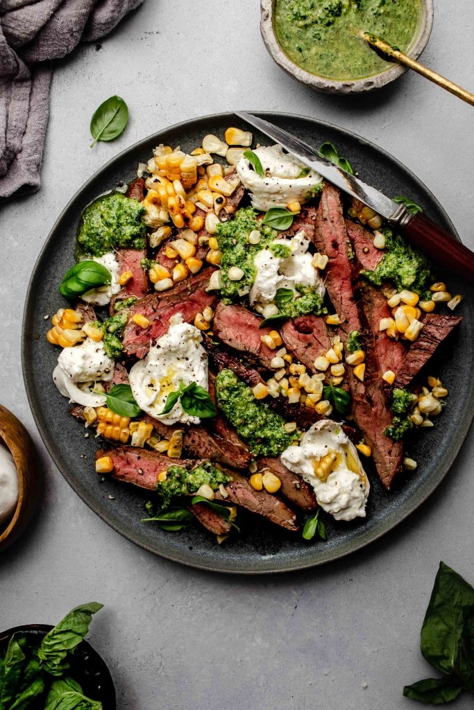 Grilled flank steak arranged on plate topped with corn, pesto and burrata cheese.