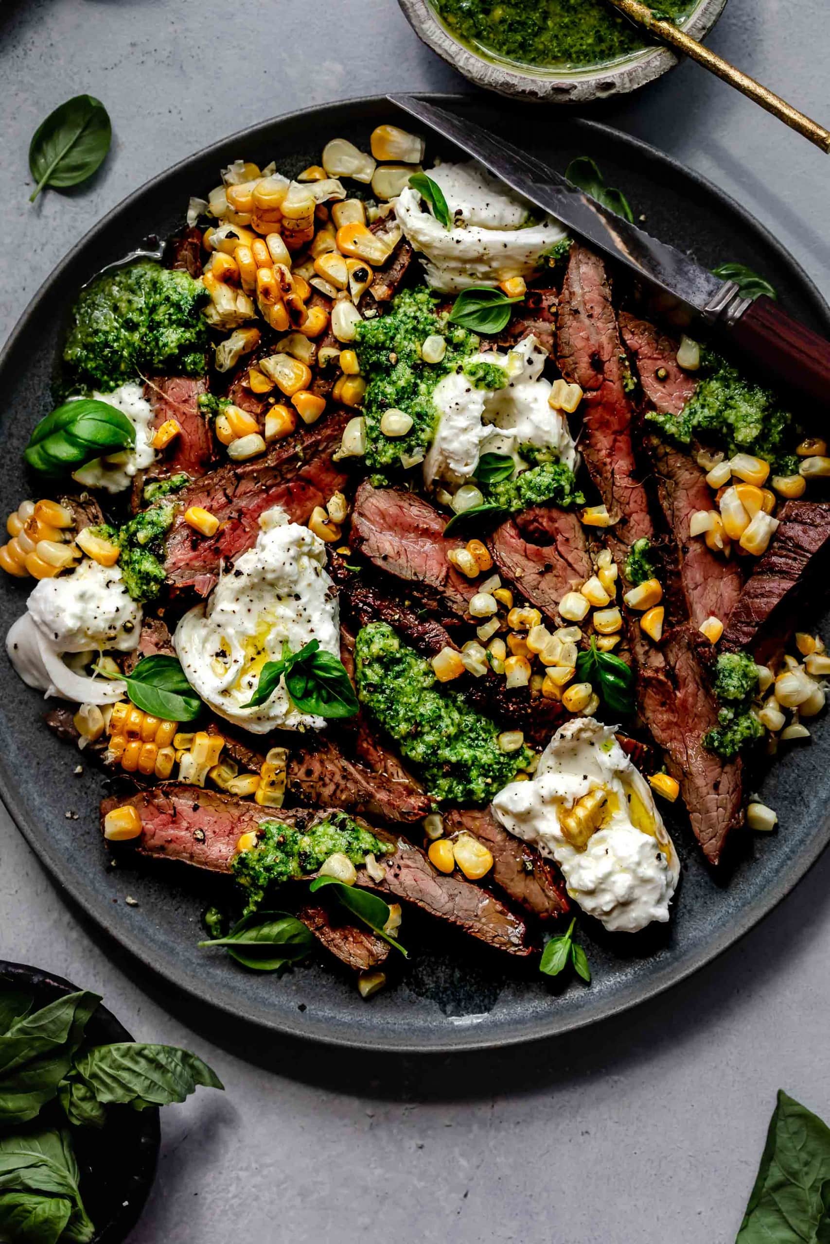 Grilled flank steak arranged on plate topped with corn, pesto and burrata cheese.
