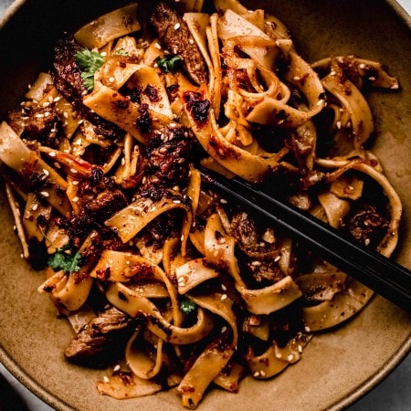 Spicy cumin lamb wide noodles in brown bowl with chopsticks.