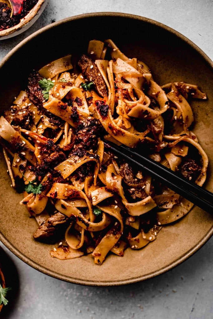 Spicy cumin lamb wide noodles in brown bowl with chopsticks.