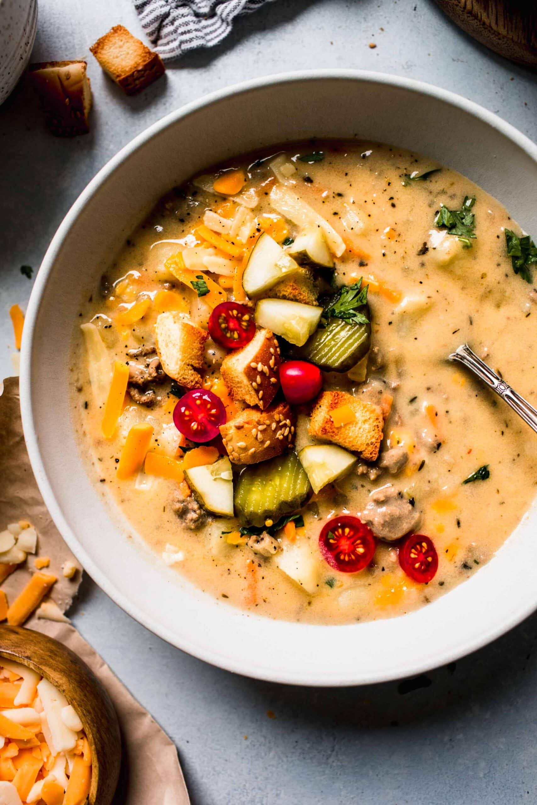 OVERHEAD CLOSE UP OF BOWL OF CHEESE BURGER SOUP TOPPED WITH PICKLES, CROUTONS AND TOMATOES.