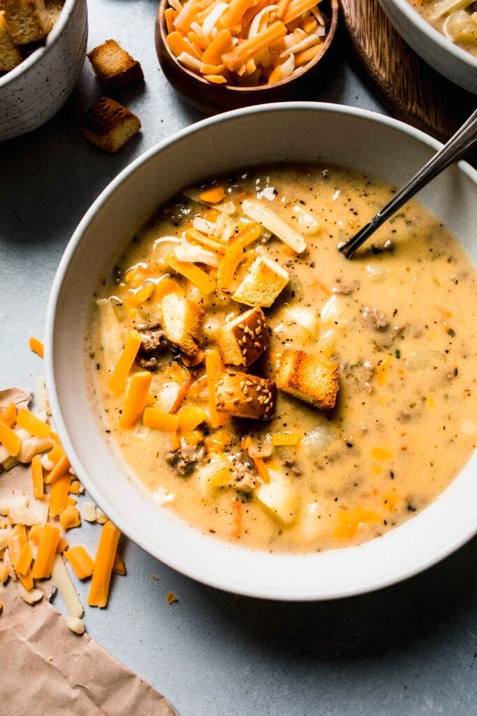 BOWL OF CHEESEBURGER SOUP IN WHITE BOWL NEXT TO BOWL OF CHEESE AND BURGER BUN CROUTONS.