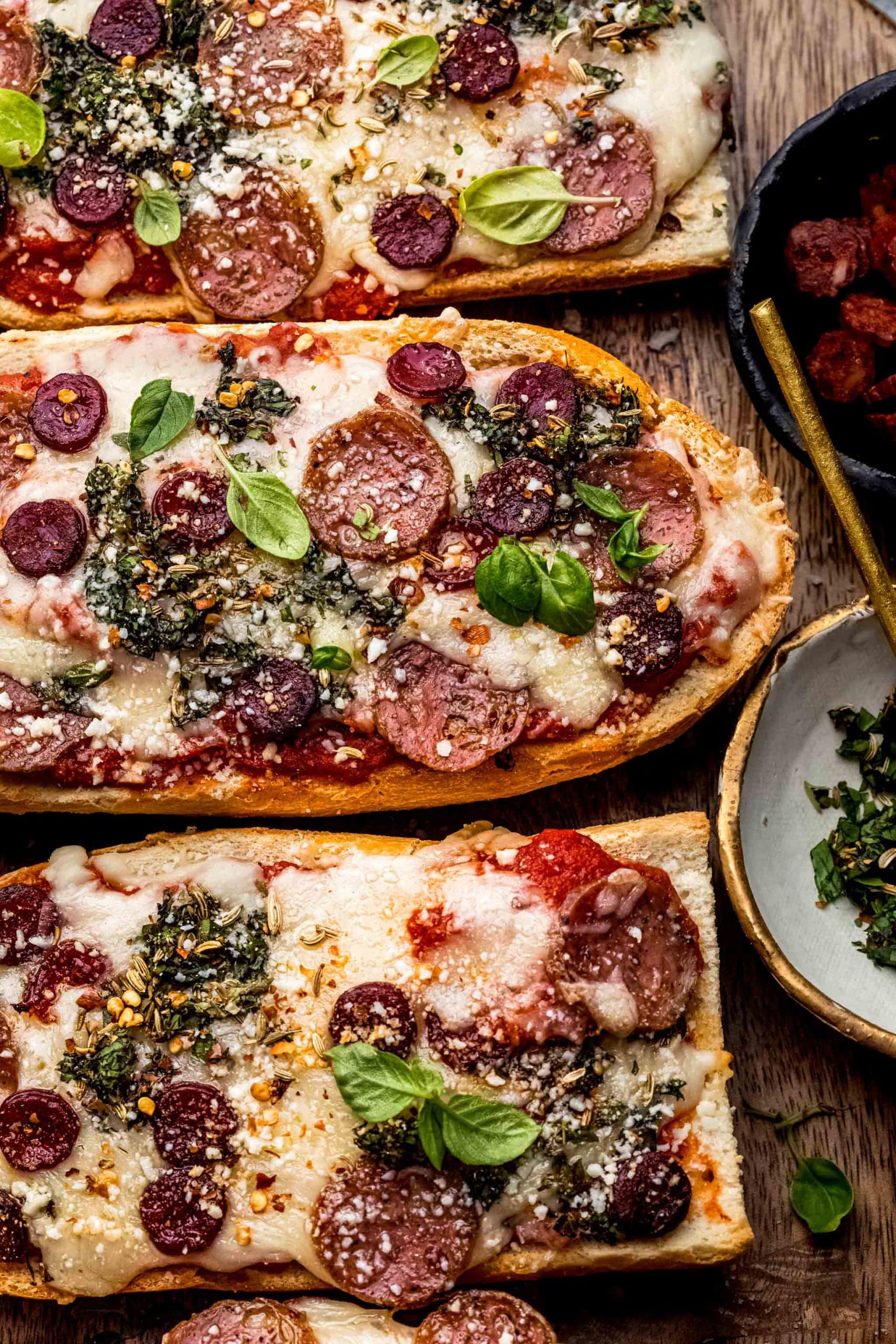 French bread pizza slices on cutting board next to small bowl of herbs and pepperoni.