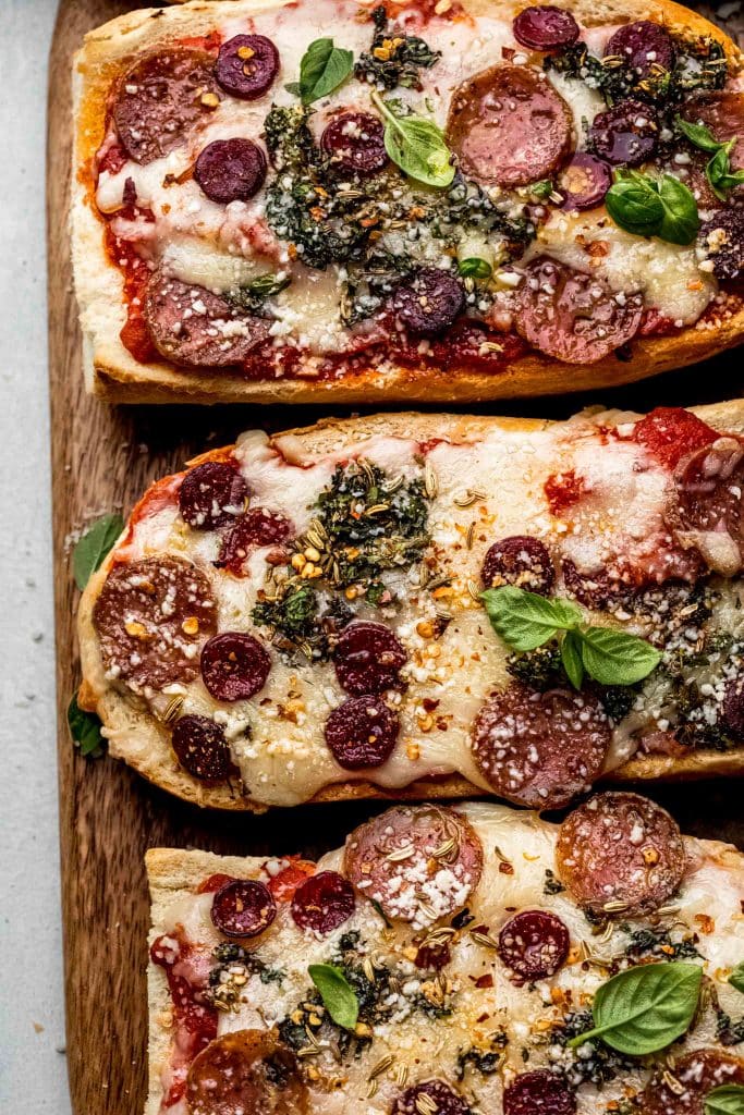 French bread pizza slices on cutting board next to small bowl of herbs and pepperoni.