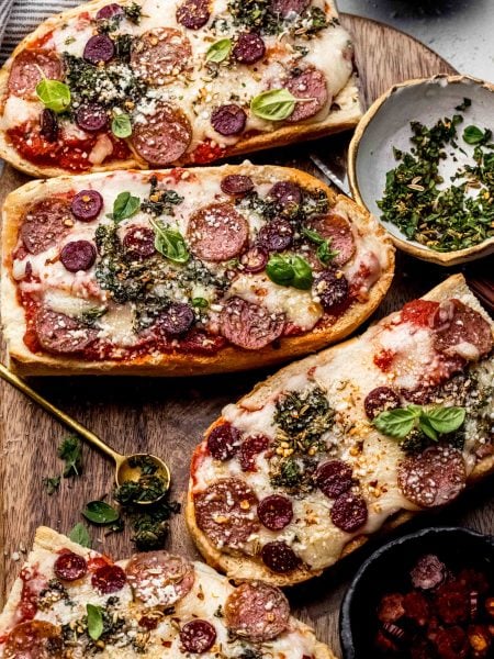 French bread pizzas on cutting board next to small bowl of herbs and pepperoni.