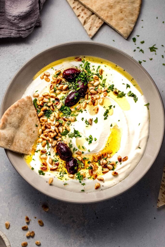 Whipped feta dip in bowl topped with olives, pine nuts and olive oil. Served with pita.