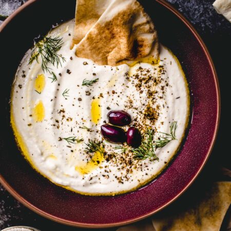 WHIPPED FETA DIP IN MAROON BOWL TOPPED WITH OLIVES, PITA BREAD AND A DRIZZLE OF OLIVE OIL.