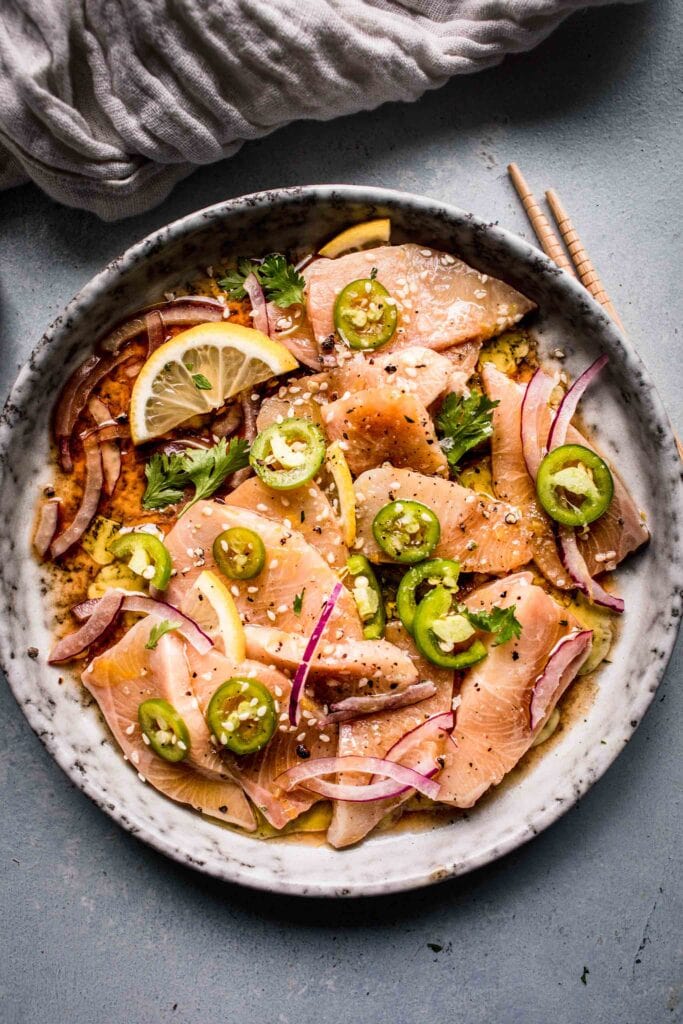 Hamachi crudo arranged on grey plate topped with lemon slices and serrano peppers.