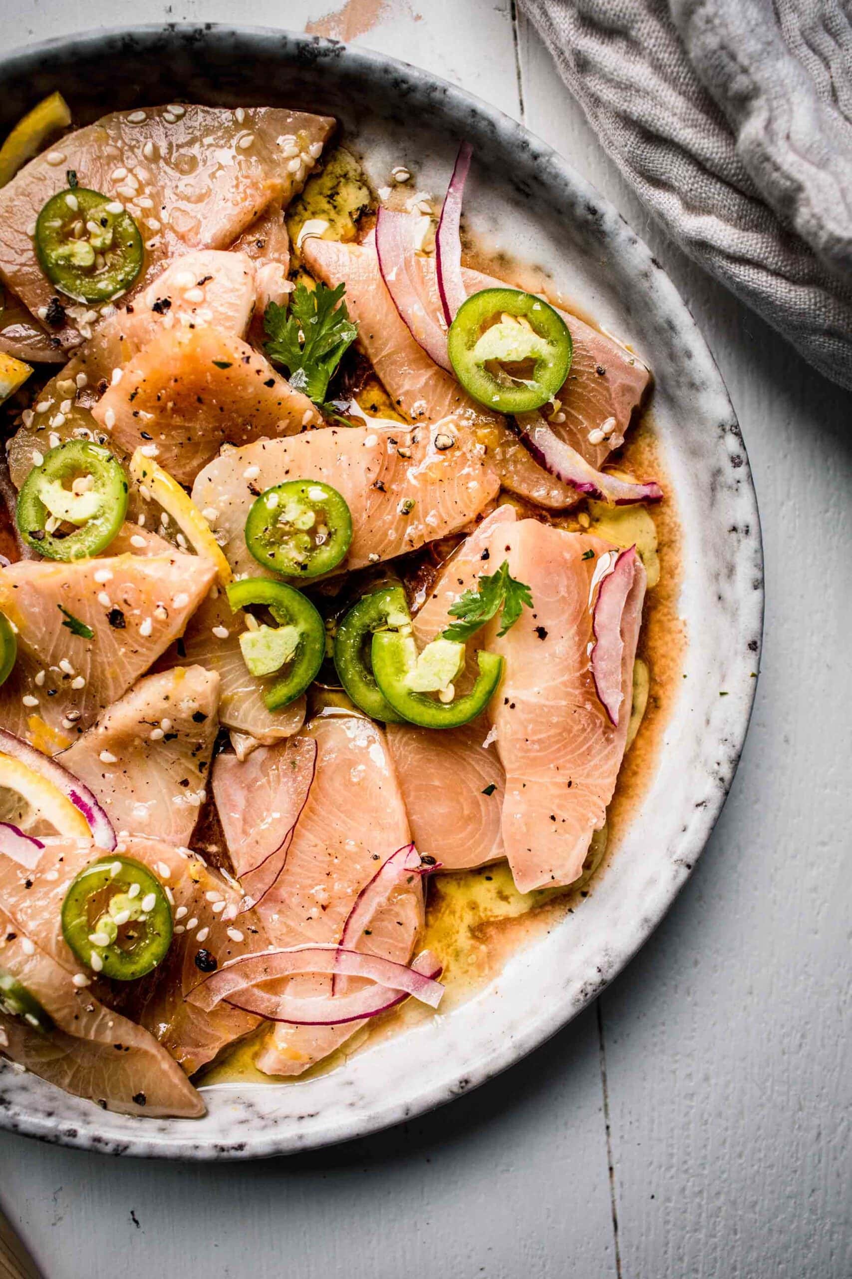 Close up shot of hamachi crudo arranged on grey plate topped with lemon slices and serrano peppers.