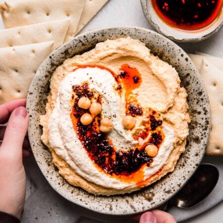 Hands holding bowl of hummus topped with whipped feta & chili oil.
