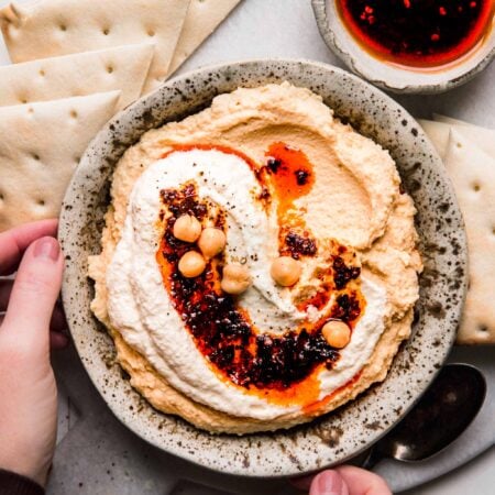 Hands holding bowl of hummus topped with whipped feta & chili oil.
