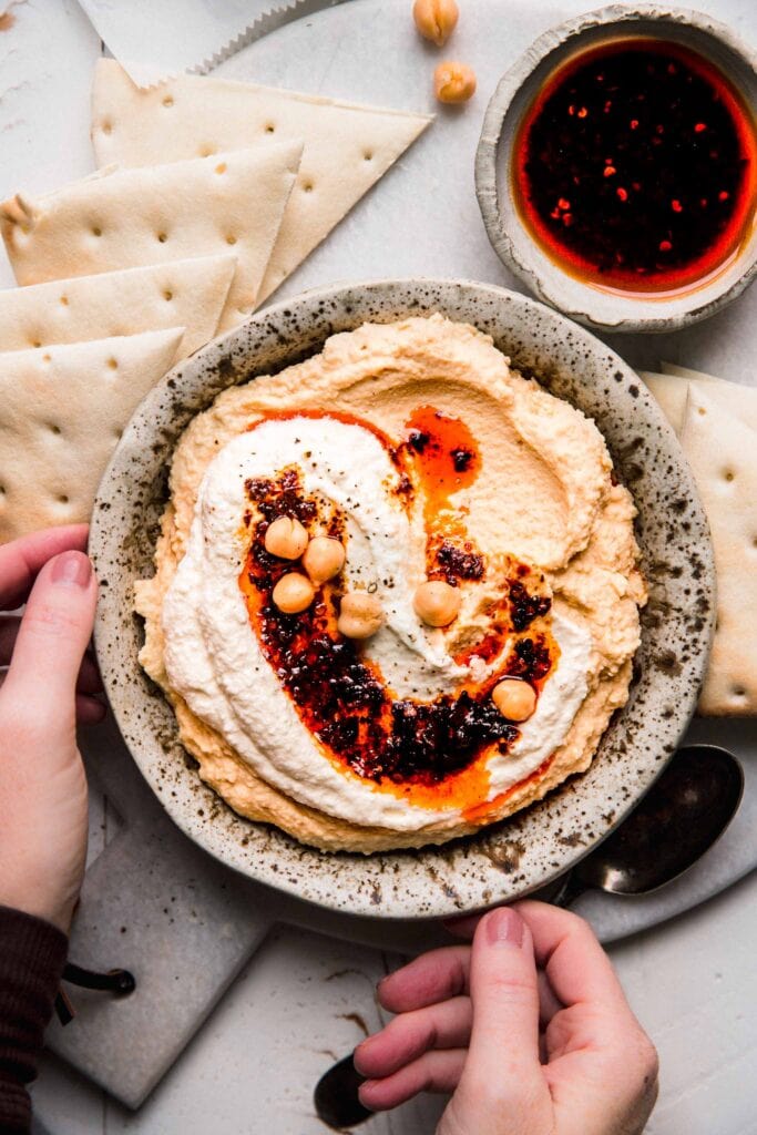 Hands holding bowl of hummus topped with whipped feta & chili oil.