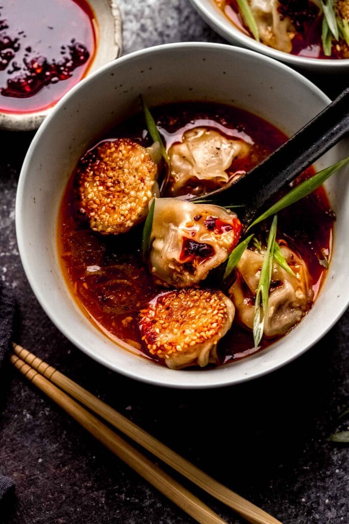 SPOON DIPPING INTO BOWL OF LAMB WONTONS TOPPED WITH CHILI OIL. 