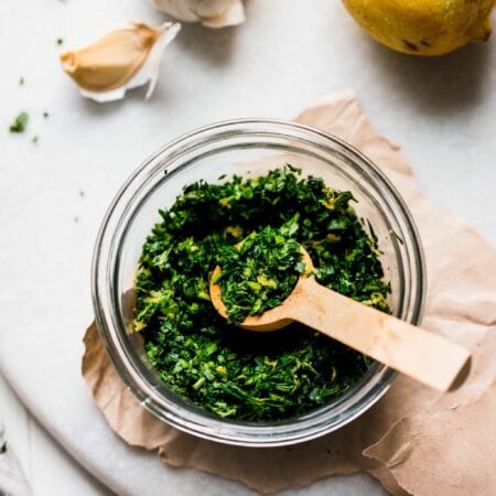 SIDE VIEW OF BOWL OF GREMOLATA NEXT TO LEMON AND GARLIC CLOVES.