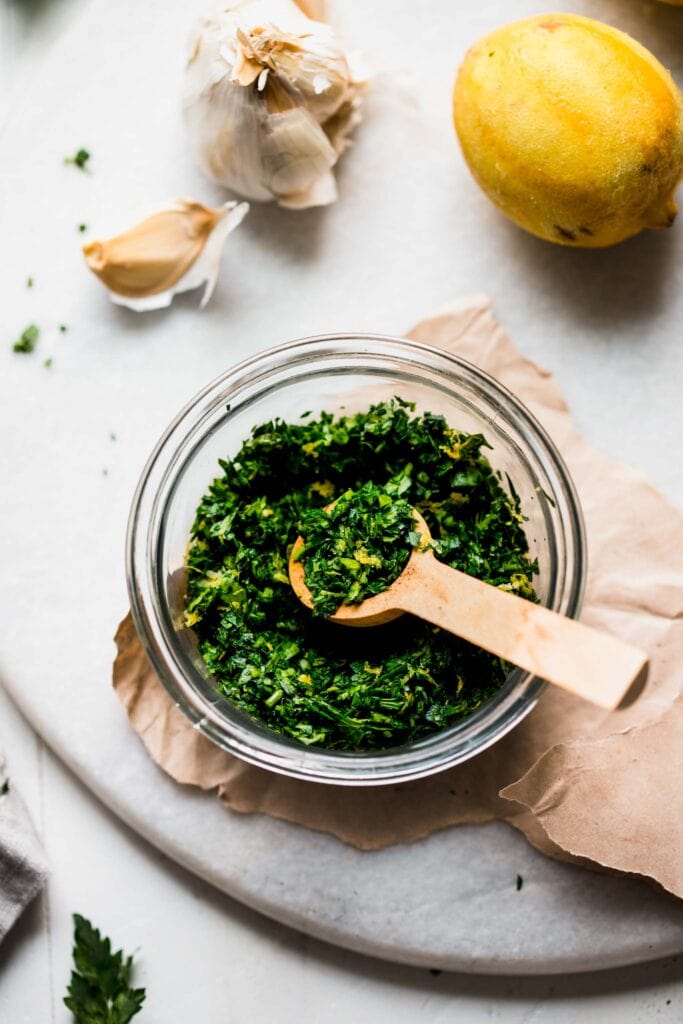 SIDE VIEW OF BOWL OF GREMOLATA NEXT TO LEMON AND GARLIC CLOVES.