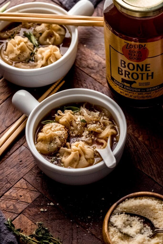 Bowls of french onion soup dumplings on wooden table, next to thyme sprigs and bowl of parmesan.