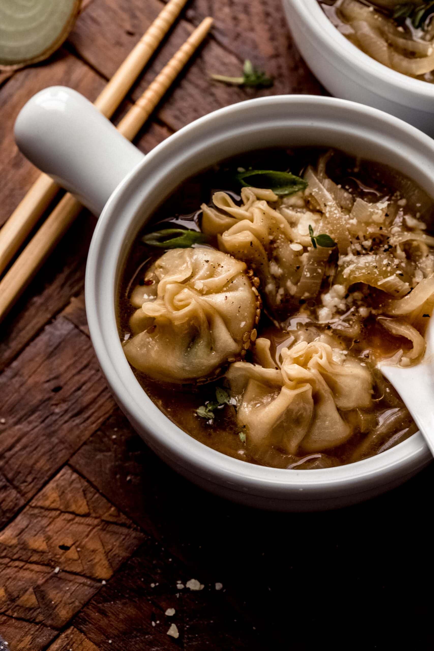 Side view of bowl of french onion soup dumplings with chopsticks in white, handled bowl.