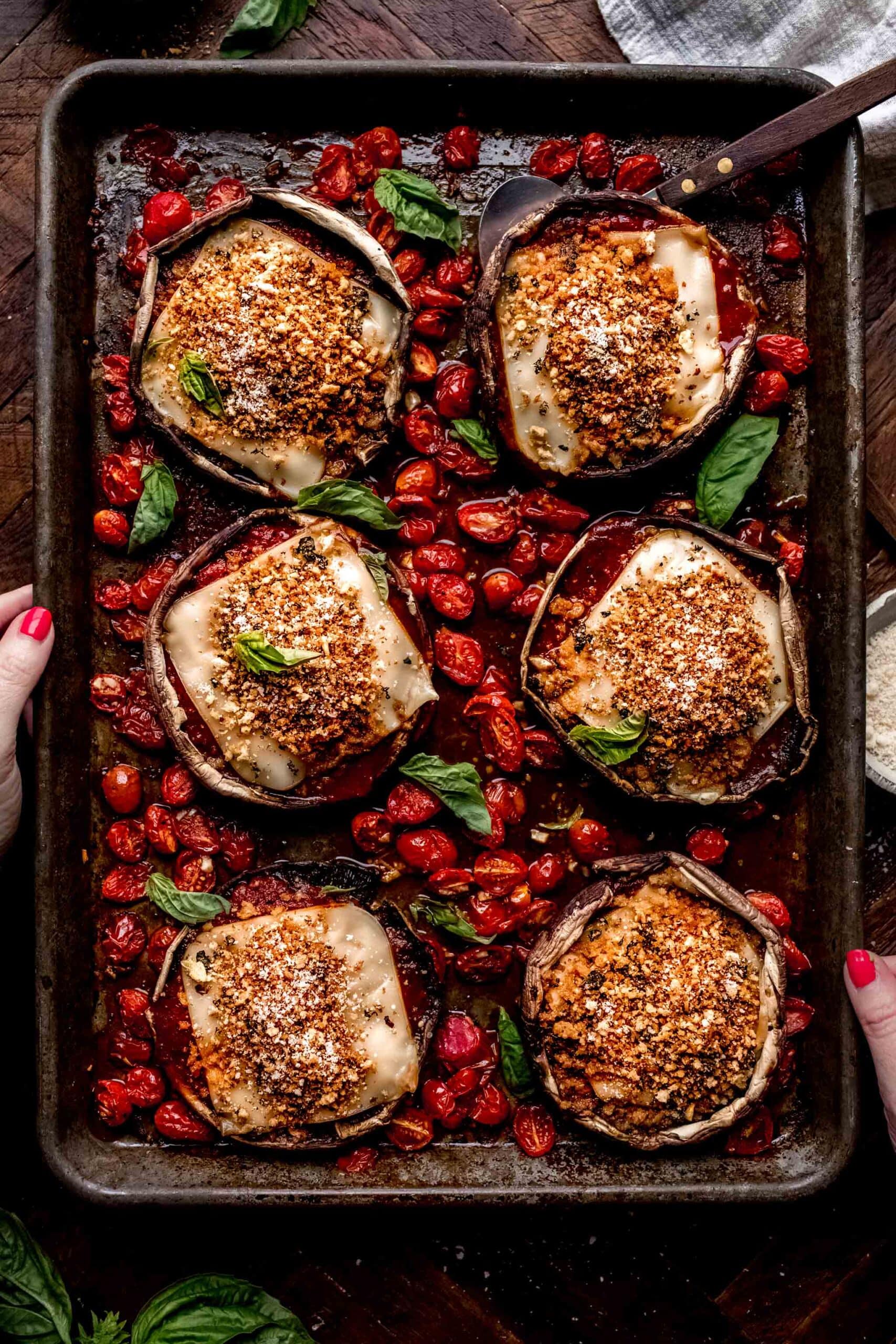 Hands holding tray of mushroom parmesan arranged on baking sheet with tomatoes scattered about.