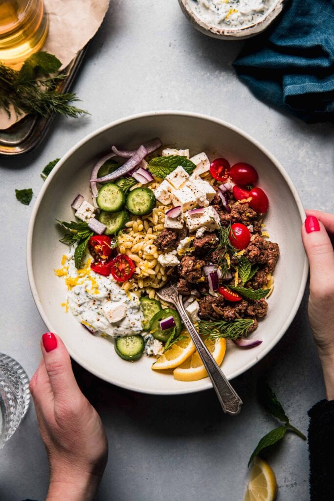 Hands holding bowl of lemon orzo with toppings. 