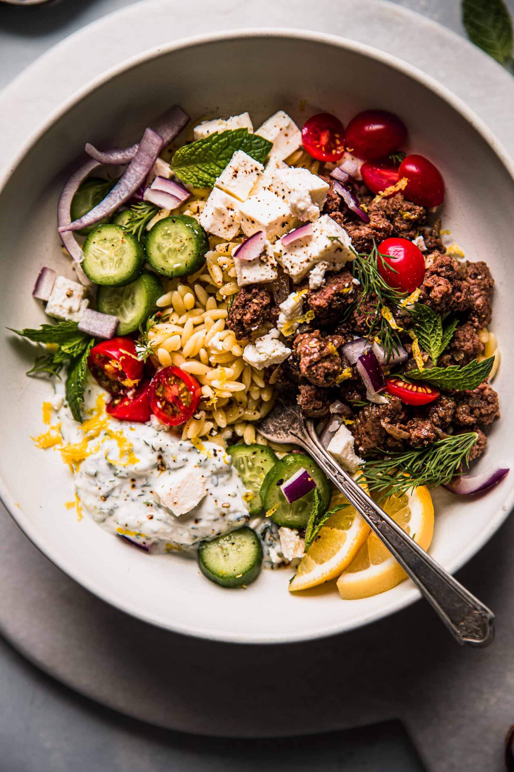 Overhead shot of lemon orzo dressed with tzatziki, cucumber slices, lemons and lamb.