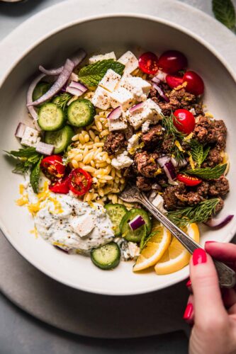 Overhead shot of lemon orzo dressed with tzatziki, cucumber slices, lemons and lamb.