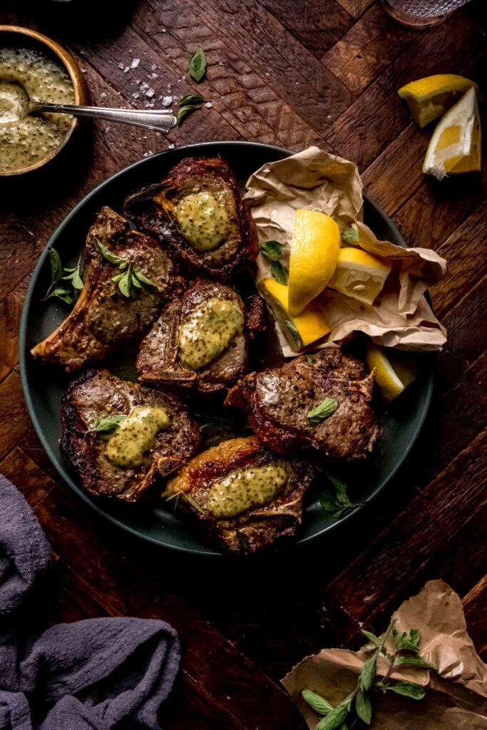 Lamb chops arranged on plate with lemon wedges, fresh herbs and dollops of mint mustard sauce. 