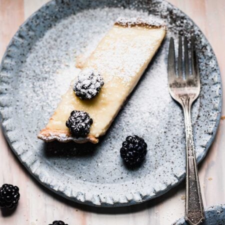 Slice of custard tart topped with powdered sugar and blackberries on blue speckled plate.