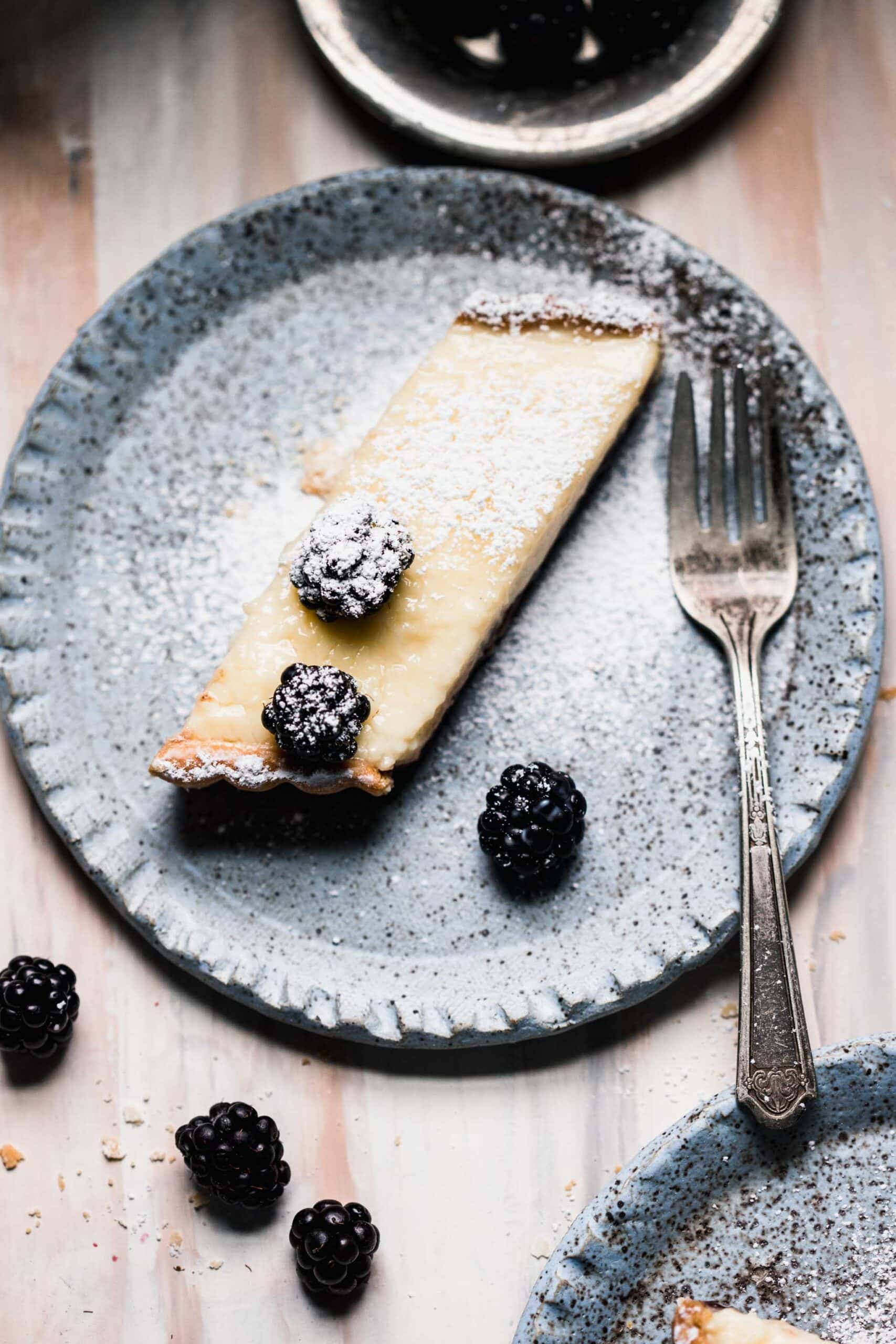 Slice of custard tart topped with powdered sugar and blackberries on blue speckled plate.
