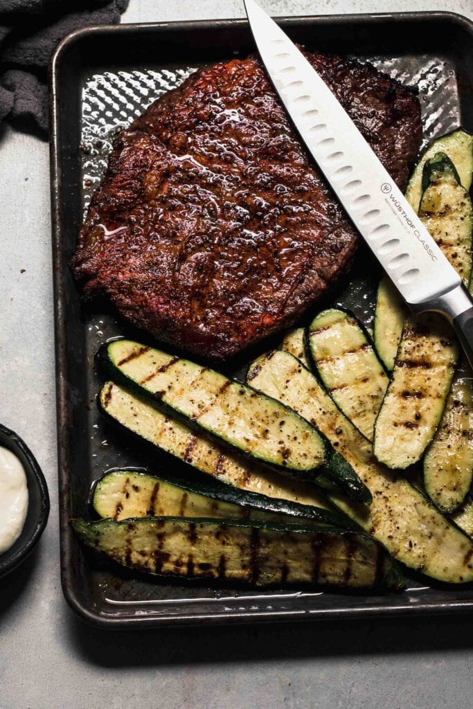 Grilled flank steak on tray next to grilled zucchini slices. 