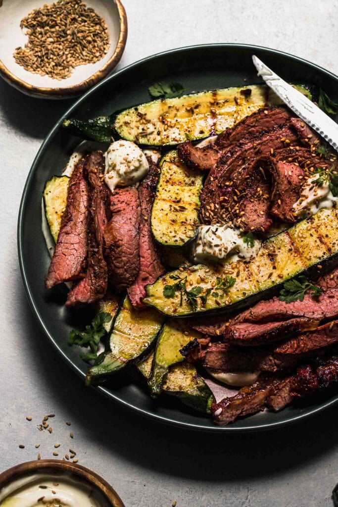 Side view of grilled zucchini and flank steak on plate next to small bowl of toasted seeds. 