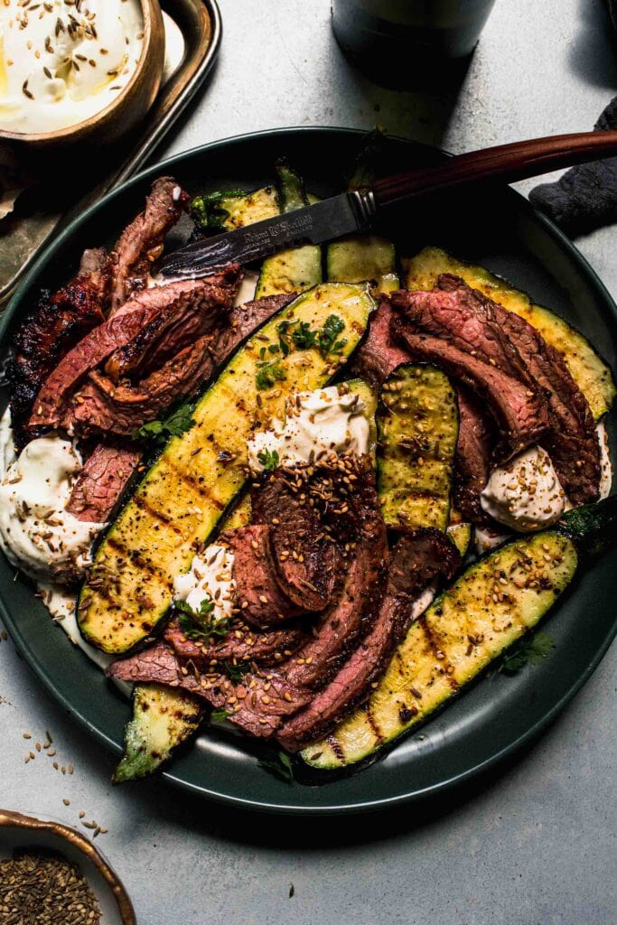 Slices of grilled steak on green plate with zucchini and feta scattered around.