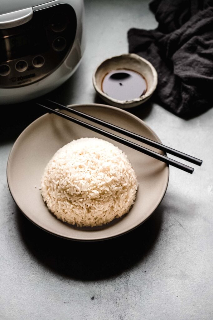 Bowl of rice with chopsticks next to rice cooker machine.