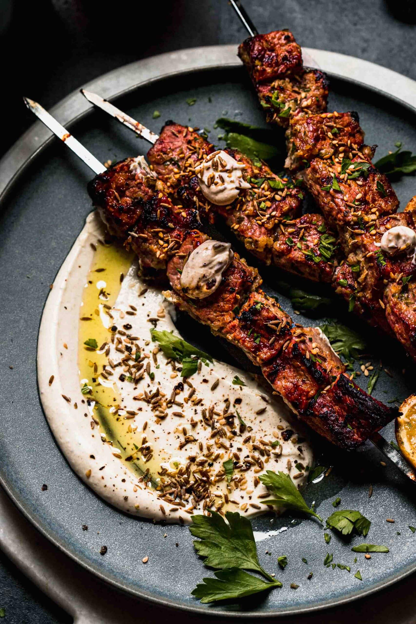 Overhead shot of lamb kebabs on plate with dollop of sesame sauce and grilled lemon.